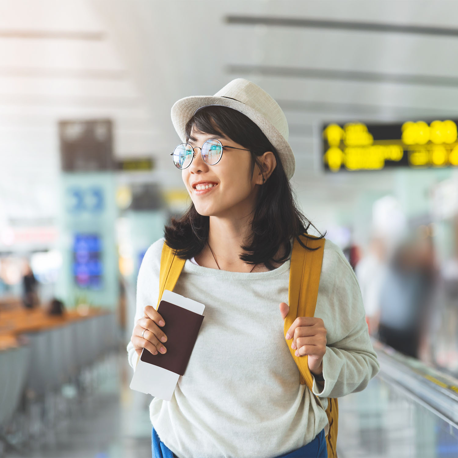Happy Asian woman wear glasses, hat with yellow backpack is holding flying ticket, passport while waiting for the flight at the hall of airport